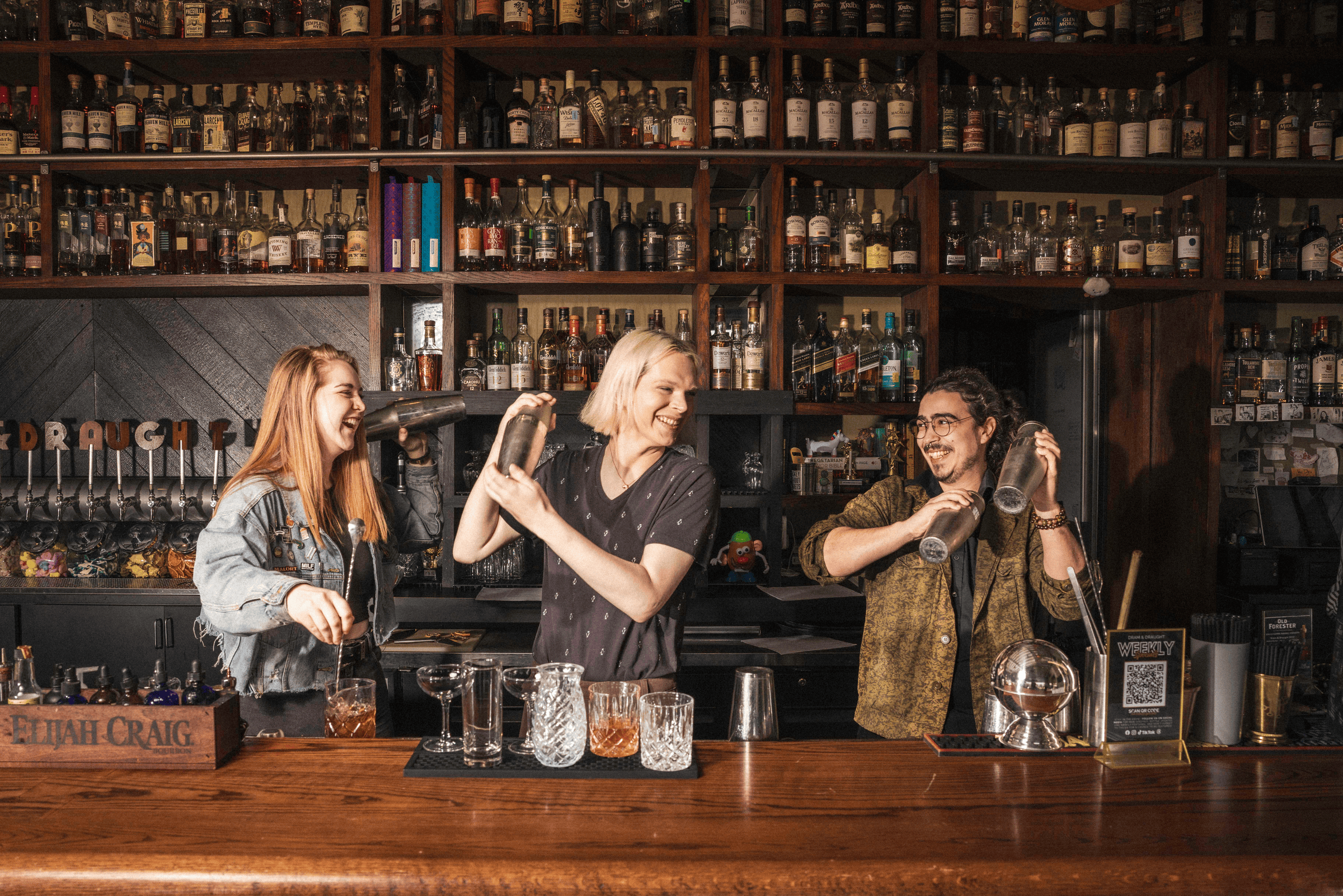 Three enthusiastic bartenders shake cocktails behind a wooden bar, surrounded by an impressive collection of liquor bottles.