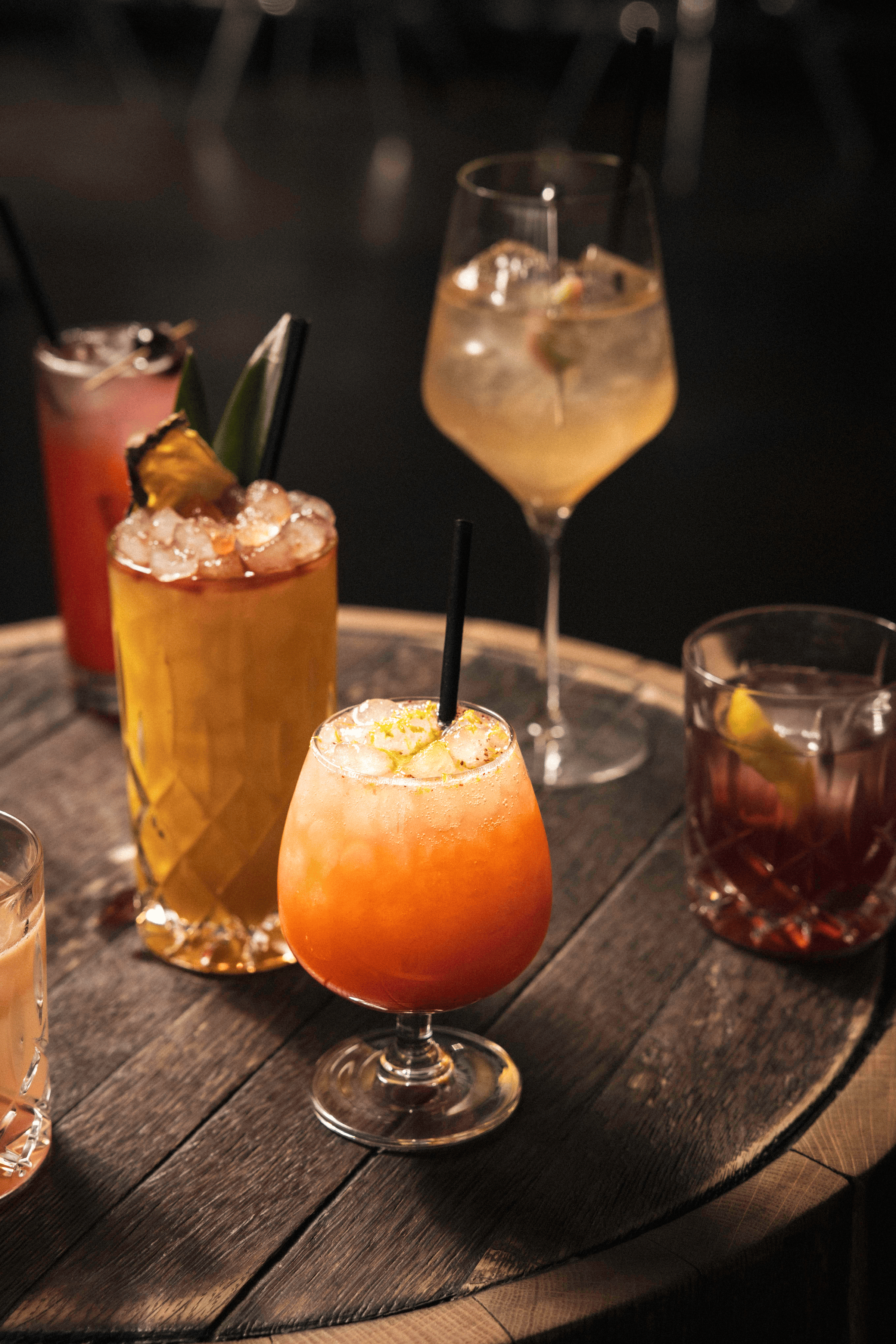 Colorful craft cocktails, glistening with ice, rest on a rustic wooden table, surrounded by soft ambient lighting.